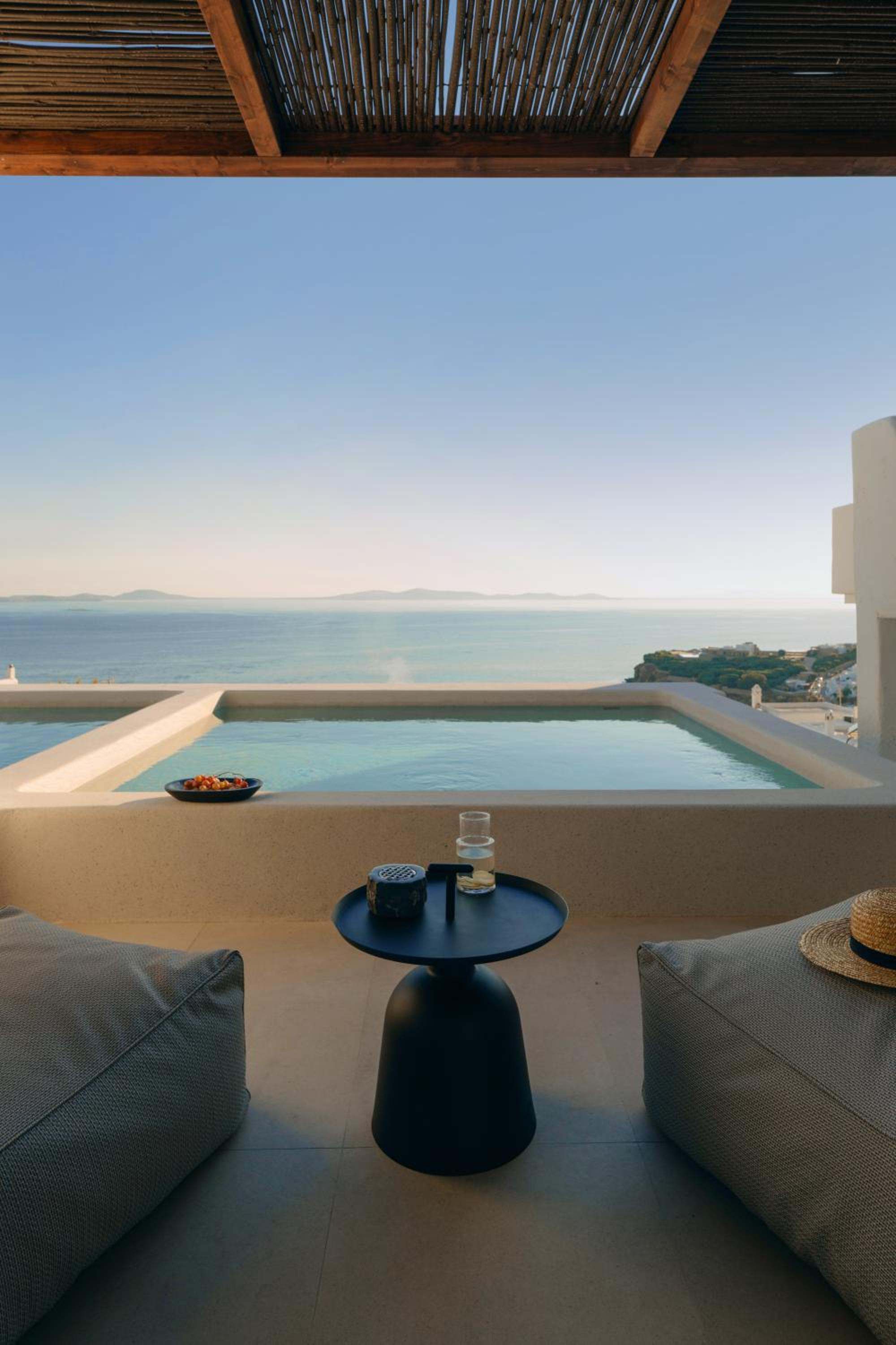 A shaded terrace with lounge cushions and a small table facing a plunge pool and the open sea