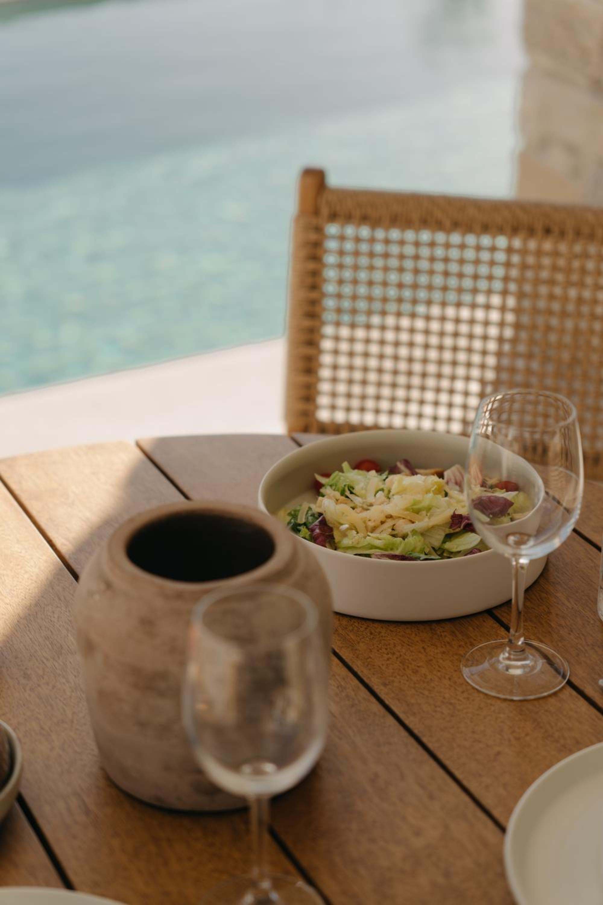 A salad and wine glasses on a timber table beside the pool